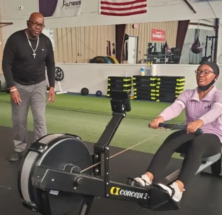a woman rows on a rowing machine as dwayne adams coaches her during the workout